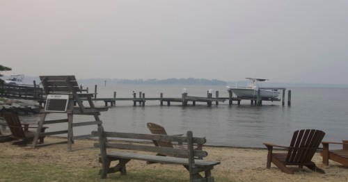 A calm waterfront scene with a dock, a boat, and wooden benches on a hazy day.