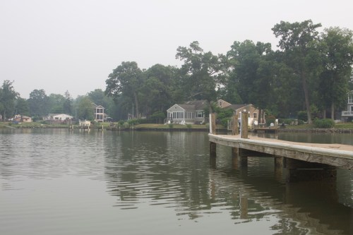 A calm lake scene with houses along the shore and a wooden dock extending into the water, surrounded by trees.