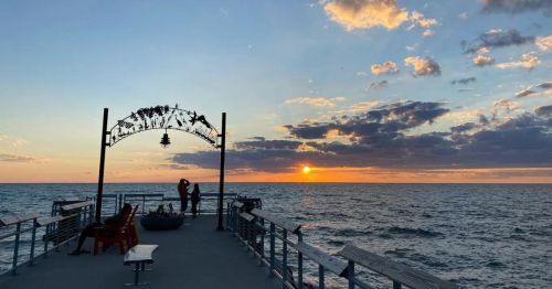 Silhouette of two people on a pier at sunset, with colorful clouds and ocean waves in the background.