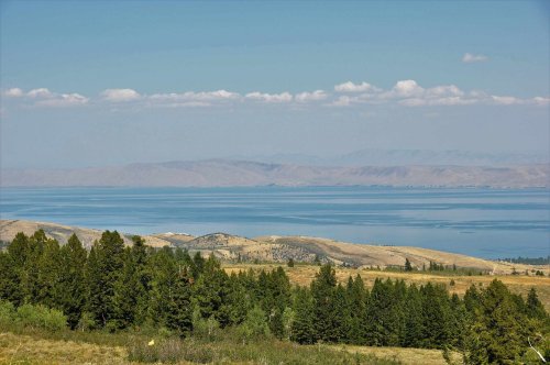 A serene landscape featuring a calm lake surrounded by rolling hills and green forests under a clear blue sky.