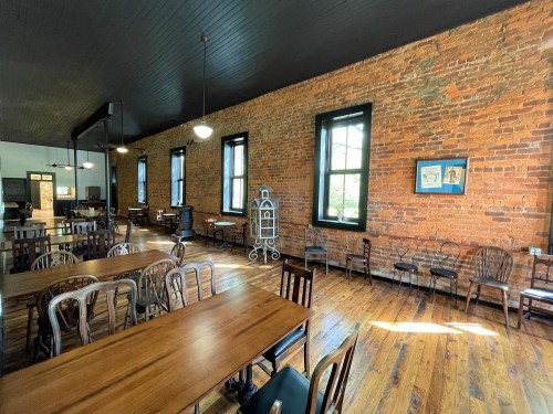 A spacious room with exposed brick walls, wooden floors, and tables surrounded by chairs, featuring large windows.