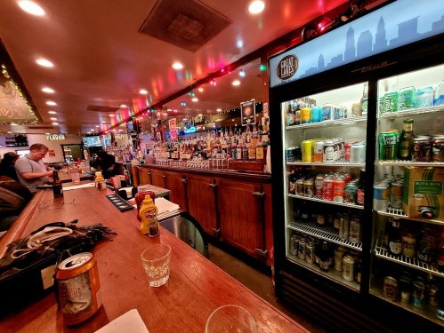 A lively bar scene with a wooden counter, shelves of liquor, and a fridge filled with various canned drinks.