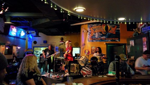 A lively band performs on stage in a bar, with colorful lights and patrons enjoying the music.