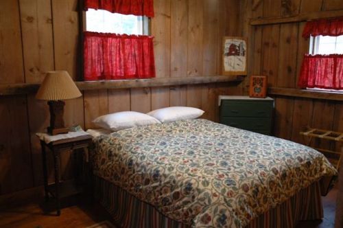 Cozy bedroom with a wooden interior, featuring a floral-patterned bedspread, two windows with red curtains, and a lamp.