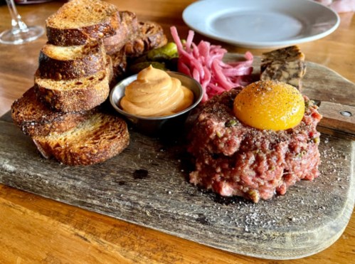 A wooden platter with toasted bread, beef tartare topped with an egg yolk, pickled onions, and a dollop of sauce.