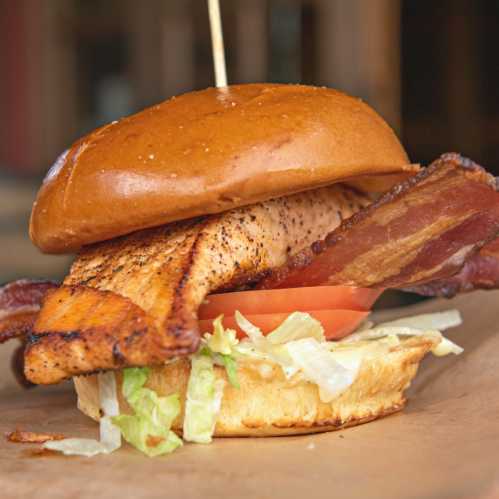 A close-up of a sandwich with a grilled salmon fillet, bacon, lettuce, and tomato on a toasted bun.