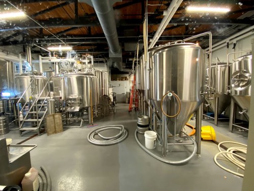 A brewery interior featuring large stainless steel fermentation tanks and hoses on the floor.