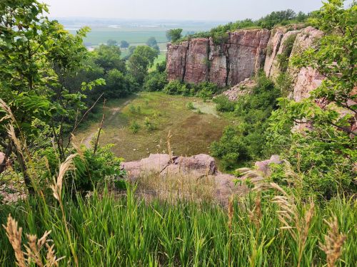 A scenic view of a rocky cliff surrounded by greenery and a grassy area below, with distant fields in the background.