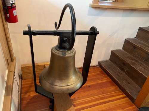 A large brass bell mounted on a wooden beam, with a staircase visible in the background.