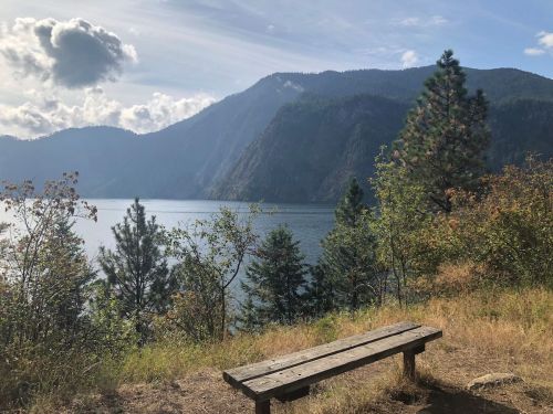 A wooden bench overlooks a serene lake surrounded by mountains and trees under a partly cloudy sky.