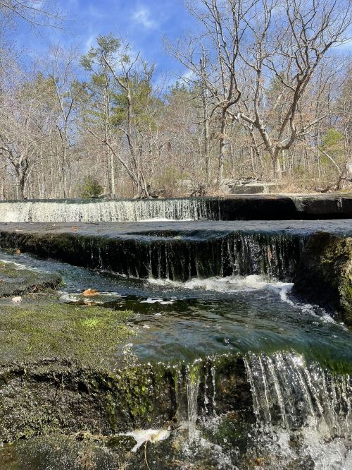 A serene waterfall cascades over rocky ledges, surrounded by bare trees and a clear blue sky.