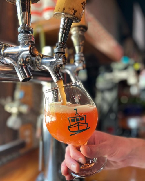 A hand pours a golden-orange beer from a tap into a glass with a boat logo.