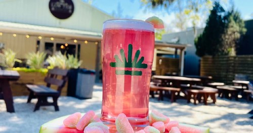 A pink drink in a glass with a logo, surrounded by candy, set outdoors with picnic tables in the background.