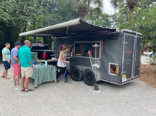 A food truck with a woman serving customers at a gravel lot, surrounded by trees and outdoor seating.