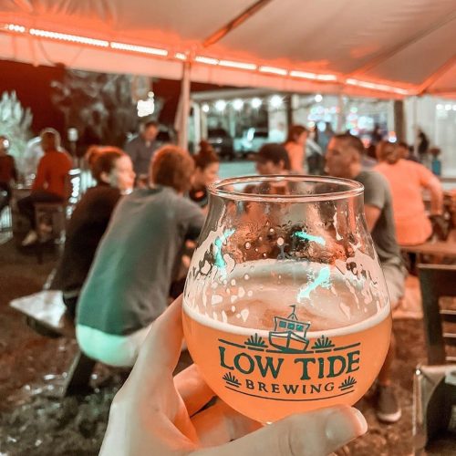 A hand holds a glass of beer with "Low Tide Brewing" logo, while people socialize under a tent in the background.