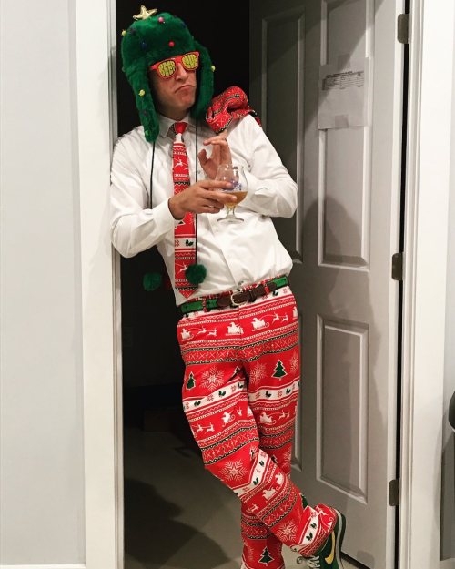 A person in a festive outfit with a Christmas tree hat, sunglasses, and holiday-themed pants poses with a drink.