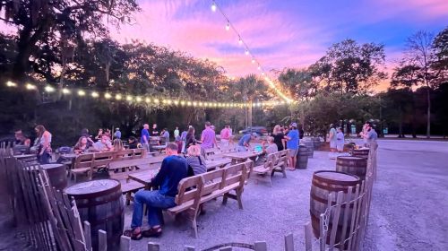 A lively outdoor gathering at sunset, with people seated at wooden tables under string lights.