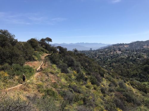 A hiker walks along a winding trail on a hillside, with mountains and a valley visible in the background under a clear blue sky.