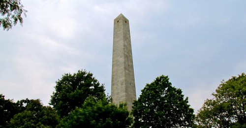 A tall stone obelisk surrounded by green trees under a cloudy sky.