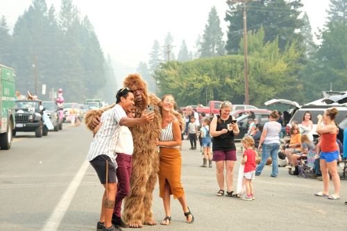 A crowd enjoys a parade, with people posing with a Bigfoot costume amidst smoke and trees in the background.