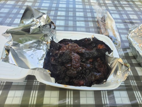 A takeout container with foil, revealing dark, charred food inside, placed on a checkered tablecloth.