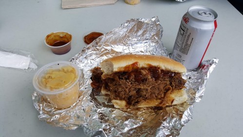 A pulled pork sandwich in foil, with barbecue sauce, a side of coleslaw, and a can of soda on a table.