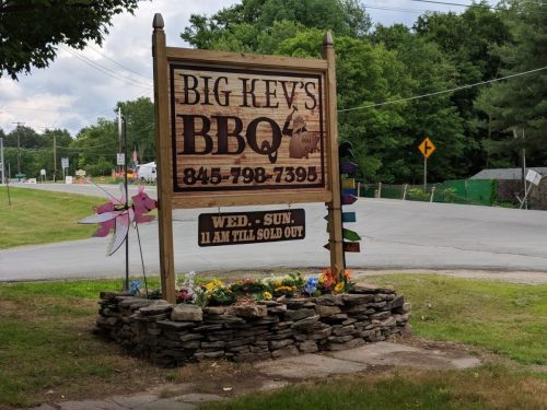 Sign for Big Kev's BBQ with a wooden design, featuring flowers and information about hours and contact number.