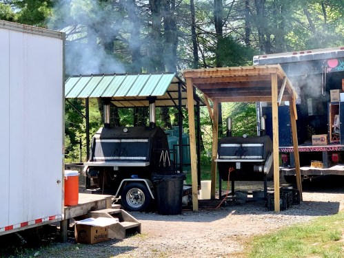 Two outdoor grills under wooden shelters, with smoke rising, surrounded by trees and parked trailers.