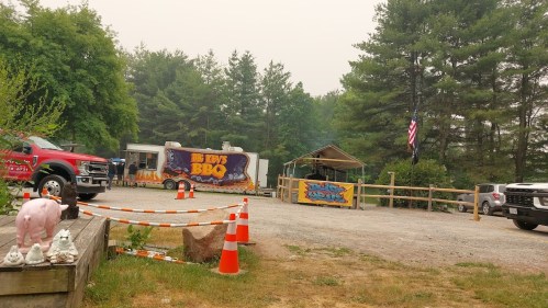 A BBQ food truck in a smoky outdoor setting, surrounded by parked cars and orange traffic cones.