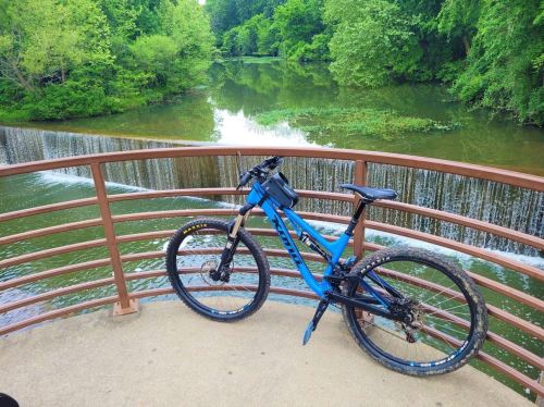 A blue mountain bike rests on a bridge overlooking a serene river with lush greenery and a small waterfall.