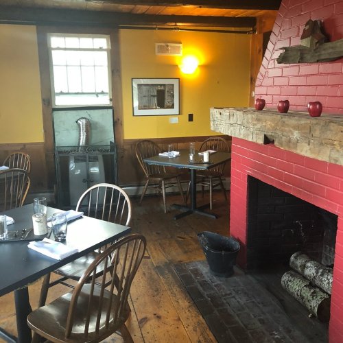 Cozy restaurant interior with wooden tables, chairs, and a red brick fireplace. Warm lighting enhances the inviting atmosphere.