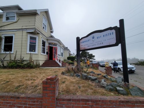 A cozy restaurant with a sign reading "Humboldt Bay Bistro," surrounded by grass and foggy weather.