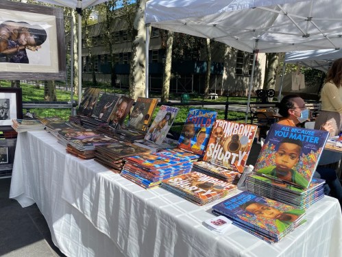 A table at an outdoor event displays colorful children's books and framed photos under a white tent.