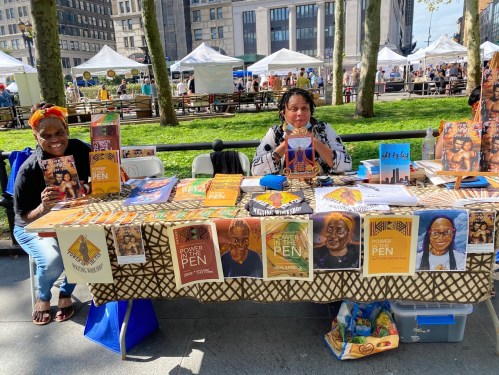 Two women at a market booth displaying books and artwork, with a vibrant table setup and tents in the background.