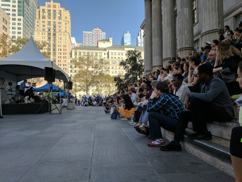 A crowd sits on steps watching a performance at an outdoor event in a city, with tall buildings in the background.