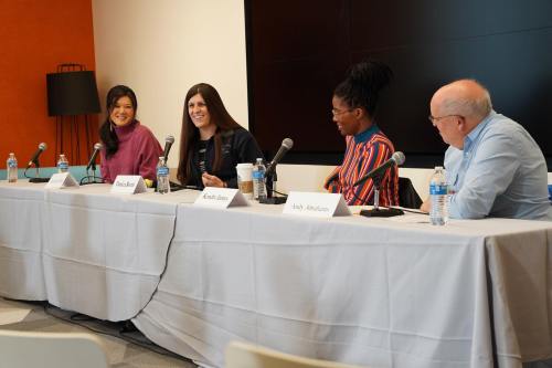 A panel discussion featuring four speakers at a table with microphones, water bottles, and a screen in the background.