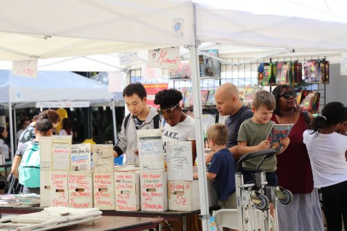A diverse group of people browsing at a market stall filled with books and comics under white tents.