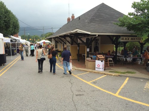 A bustling outdoor market with vendors and visitors near a historic building under a cloudy sky.