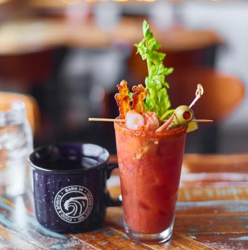 A vibrant Bloody Mary garnished with bacon, celery, and olives, alongside a black coffee mug on a wooden table.