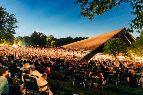 A large crowd gathers at an outdoor concert venue, surrounded by trees, under a clear evening sky.