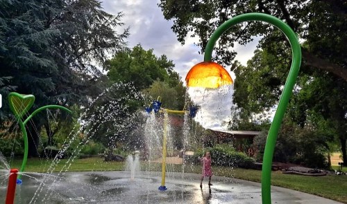 A child plays in a colorful splash pad with water features under a cloudy sky. Green trees surround the area.