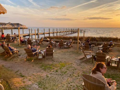 People relax in Adirondack chairs by the water, enjoying a sunset view over the pier and calm sea.