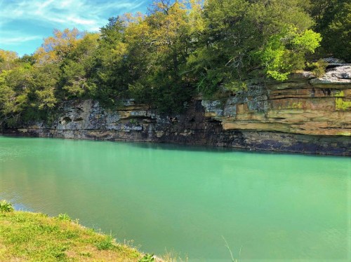 A serene river with turquoise water flows beside a rocky, green-covered cliff under a clear blue sky.