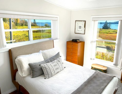 Cozy bedroom with a bed, decorative pillows, and large windows showcasing a scenic view of a lake and autumn foliage.