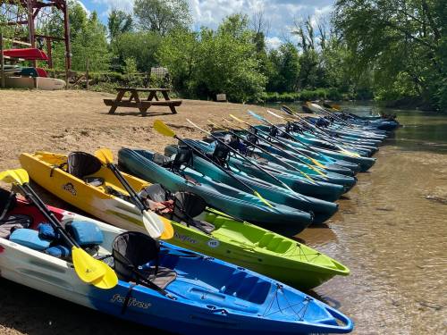 A row of colorful kayaks lined up on a sandy shore by a calm river, surrounded by greenery and blue skies.