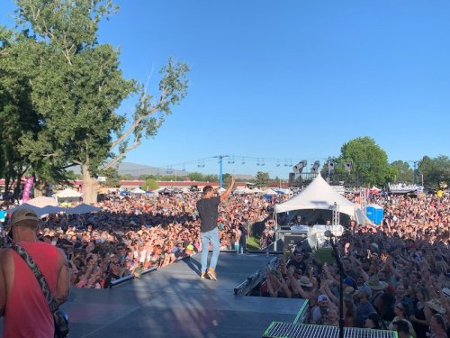 A performer on stage gestures to a large crowd at an outdoor music festival under a clear blue sky.