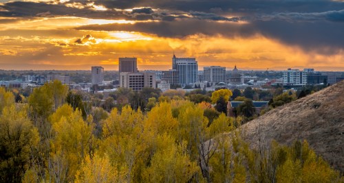 A city skyline at sunset, surrounded by autumn trees, with dramatic clouds and warm colors in the sky.