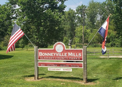 Sign for Bonneyville Mills in Elkhart County Parks, with American and state flags, open May to October, Wednesday to Sunday.