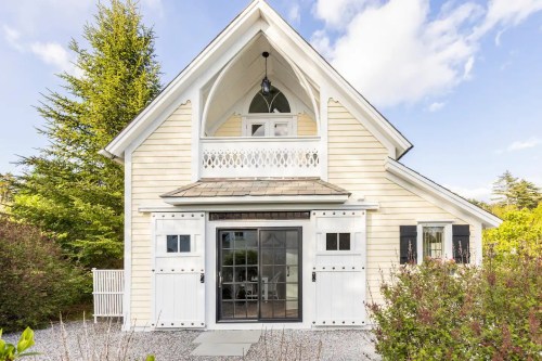 Charming light-colored house with a balcony, large windows, and a landscaped garden under a blue sky.