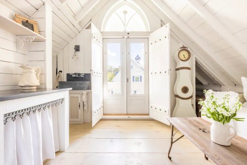 Bright, airy attic room with white walls, open double doors, and a rustic wooden table with flowers.